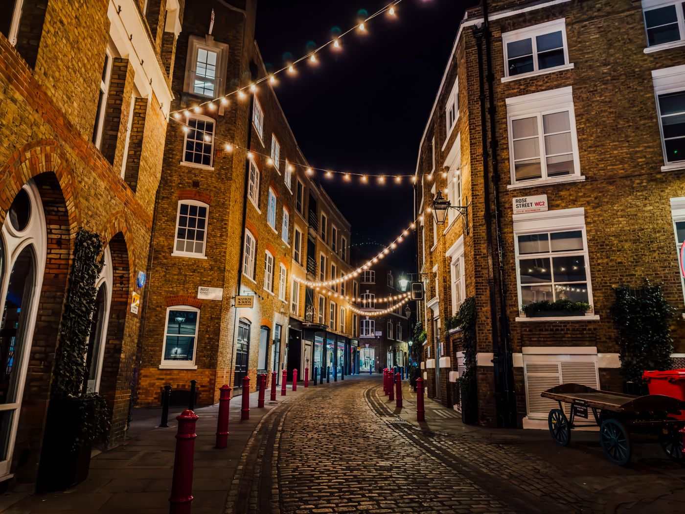Illuminated cobblestone street in Soho, London with string lights and historic buildings at night.