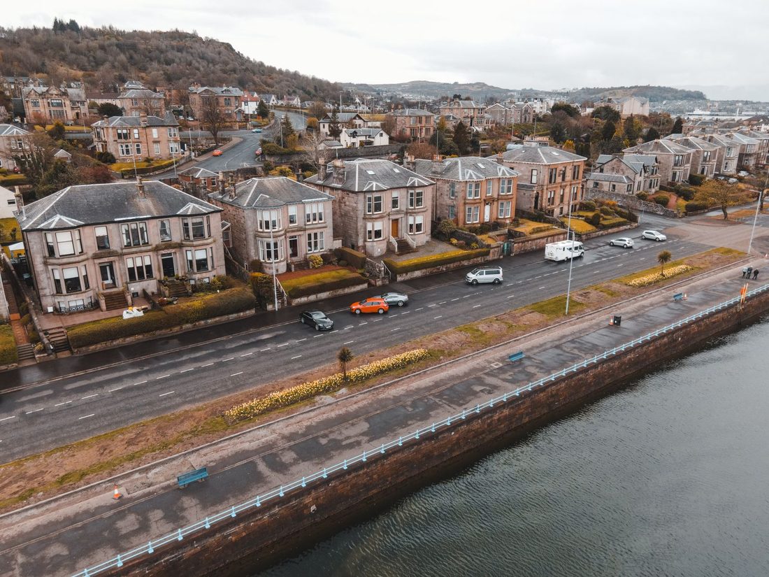 A scenic aerial view of Greenock, Scotland featuring Victorian homes along a coastal road with visible traffic.