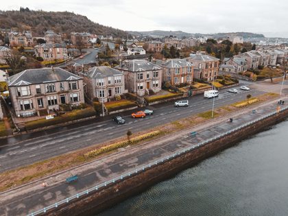 A scenic aerial view of Greenock, Scotland featuring Victorian homes along a coastal road with visible traffic.
